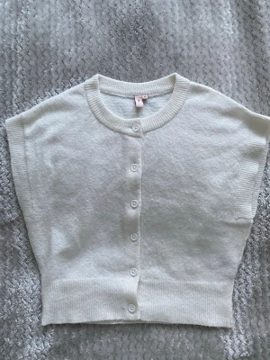 Vit ärmlös kofta från Nelly XS - Supersöt vit ärmlös kofta från Nelly i storlek XS. Koftan har rund halsringning, ribbade kanter och knäppning med fem vita knappar framtill. Perfekt att styla över en topp eller klänning för en clean look.