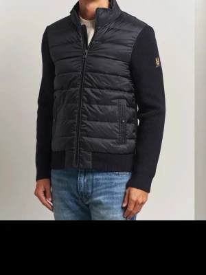 Belstaff cardigan - Säljer en svart pufferjacka med stickade ärmar och hög krage. Jackan har dragkedja framtill och två fickor med knappdetaljer. Perfekt för dig som vill ha en modern och stilren look med en twist av stickat material på ärmarna. Kvitto finns inte mycke använd