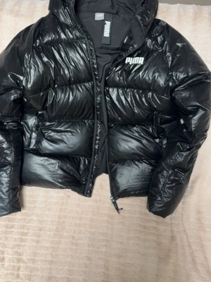Svart pufferjacka från Puma - Säljer en svart pufferjacka från Puma med glansig finish och huva. Jackan har dragkedja framtill och Puma-logga på bröstet. Perfekt för kalla dagar och riktigt snygg till streetwear-looken. Storlek L men det passar M oxå 