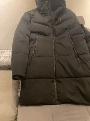 Svart lång vinter pufferjacka från Zara M herr - Säljer en svart lång pufferjacka från Zara med huva och stora dragkedjor framtill. Jackan har två snedställda fickor med dragkedja och är vadderad för extra värme. Perfekt för kalla dagar och riktigt snygg till vintern.