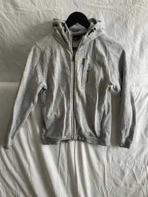 Grå sail racing tröja -  ⸻  Grå zip-hoodie i mjukt och bekvämt material. Tröjan har hel dragkedja framtill, huva med justerbara dragsnören samt två sidfickor och en bröstficka med dragkedja. Diskret tryck/branding på bröstet och liten logotyp på huvan baktill. Klassisk passform som passar både till vardag och fritid. Använd men i gott skick. 