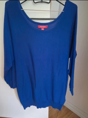 Blå långärmad tröja från Guess - Snygg blå långärmad tröja från Guess med rund hals och guldfärgade knappar på ärmsluten. Tröjan har ribbade muddar och är tillverkad i mjuk bomull, perfekt för dig som gillar stilrena och enkla plagg med en liten detalj.