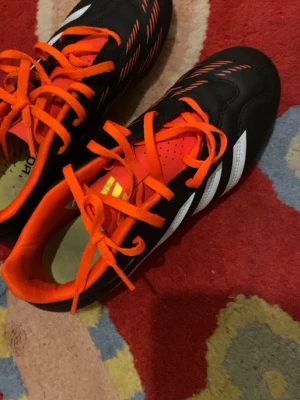 Adidas Predator fotbollsskor svart/orange - Säljer ett par Adidas Predator fotbollsskor i svart med vita ränder och orange detaljer. Skorna har snörning, tålig syntetöverdel och dobbar för bra grepp på planen. Insidan är fodrad i orange och designen är riktigt sportig.