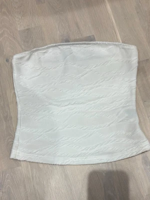 Vit strukturerad tubtopp - Säljer en stilren vit tubtopp med diskret strukturerat mönster. Toppen är axelbandslös och har en lätt figurnära passform. Perfekt att matcha med jeans eller kjol för en clean look.