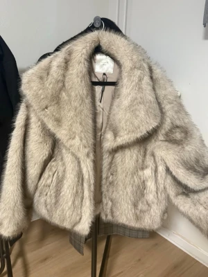 Beige pälsjacka från Neo Noir - Säljer en fluffig och lyxig beige pälsjacka från Neo Noir med bred krage och långärmad design. Jackan har en snygg croppad passform och är fodrad på insidan för extra komfort. Perfekt för dig som vill ha en statement piece i garderoben. Använd 3 gånger och inga defekter, ser helt ny ut