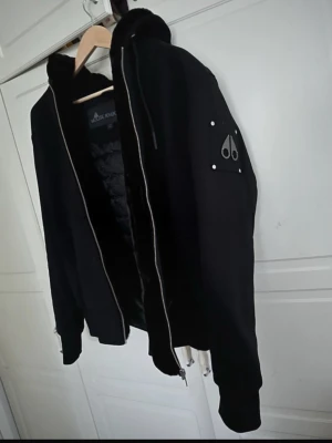 Svart pufferjacka från Moose Knuckles - Säljer en svart pufferjacka från Moose Knuckles med huva och dubbla dragkedjor. Jackan har en snygg logga på ärmen och är fodrad för extra värme. Perfekt för kalla dagar och har en modern, clean look.