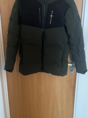 Grön pufferjacka från Sail Racing  - Säljer en mörkgrön pufferjacka från Sail Racing med svarta partier upptill och dragkedjor framtill. Jackan har huva, lång ärm och en clean, sportig look. Perfekt för kalla dagar och riktigt snygg till streetwear-stilen. Storleken är 170, men funkar som XS. 