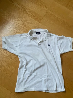 Vit pikétröja från Fred Perry - Klassisk vit pikétröja från Fred Perry i 100% bomull med broderad logga på bröstet. Tröjan har korta ärmar, krage och knappar framtill. Perfekt till en clean och sportig stil.