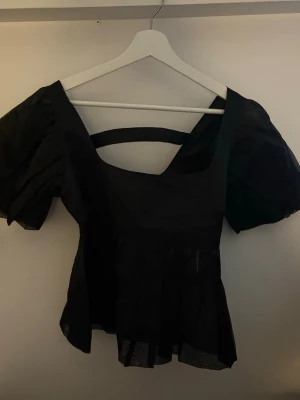 Svart blus med puffärm från BikBok - Supersnygg svart blus från BikBok med puffiga korta ärmar och fyrkantig ringning. Blusen har en volangig nederdel och en cool öppen detalj i ryggen. Perfekt att styla med jeans eller kjol för en trendig look.