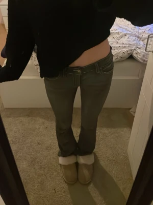 Grå bootcut jeans med vit kontrastsöm - Snygga grå jeans med låg midja och bootcut-snitt. De har vita kontrastsömmar och en unik bakficka med lock och knappdetalj. Materialet är klassiskt jeans och passformen är normal. Perfekt för dig som gillar detaljer och vill sticka ut lite extra.