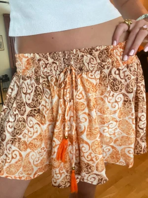 Mönstrade orange loose kjol/shorts med snörning - Säljer en flowig kjol/shorts med orange och brunt mönster, bred resår i midjan och snörning med tofsar framtill. De har en loose fit och är perfekta för varma dagar. Materialet känns lätt och luftigt, troligen bomull eller viskos. Storleken är S men resårbandet i midjan är väldigt stretchig så kan passa på M också. 