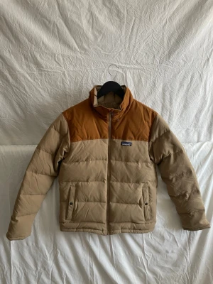 Beige och brun pufferjacka Patagonia - Säljer en snygg pufferjacka från Patagonia i beige med bruna detaljer över axlarna. Jackan har hög krage, dragkedja framtill och två fickor med knappar. Perfekt för kyliga dagar och riktigt skön att bära.
