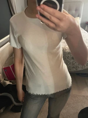 Vit ribbad tröja med vågig kant - Vit ribbad tröja i mjukt och stretchigt material. Tröjan har kort ärm samt dekorativa vågiga kanter vid ärmslut och nederkant, vilket ger ett feminint och stilrent uttryck. Enkel att matcha med jeans, kjol eller under en kofta. Passar perfekt till både vardag och mer uppklädda tillfällen.