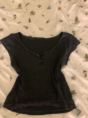 Randig topp från Brandy Melville - Kortärmad topp från Brandy Melville i mörkgrått med tunna ljusgrå ränder. Modellen har en v-ringning med knappdetalj framtill. Brandt mellville är onezine men skulle säga att den passar s-m (även xs går om man vill ha mer oversized). 
