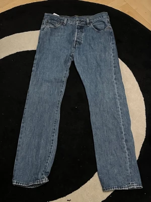 Levi's 501 blå jeans W30 L30 - Klassiska blå Levi's 501 jeans med rak passform och femficksdesign. Jeansen har knappgylf och är tillverkade i slitstarkt denimtyg av bomull. Perfekta för dig som gillar en tidlös och avslappnad look. 