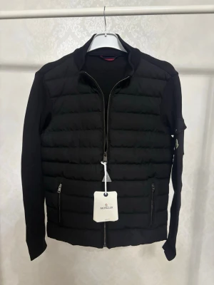 Moncler Cardigan - Hej! Säljer en svart Cardigan från Moncler med quiltad front och släta ärmar. Jackan har dragkedja framtill, två sidofickor med dragkedja och Moncler-logga på ärmen. Insidan har den klassiska serietidningsmotivet. Perfekt för dig som vill ha en stilren och modern look året runt 😊❄️