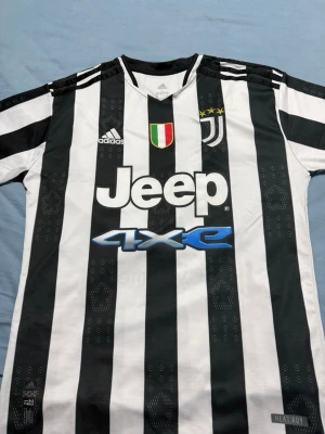 Juventus randig fotbollströja Adidas - Juventus officiella fotbollströja från Adidas med klassiska svarta och vita ränder, broderade klubbmärke och tre stjärnor på bröstet. Tröjan har Jeep och 4XE som sponsortryck samt Cygames på ryggen. Tillverkad i lätt polyester med HEAT.RDY-teknologi för extra komfort.