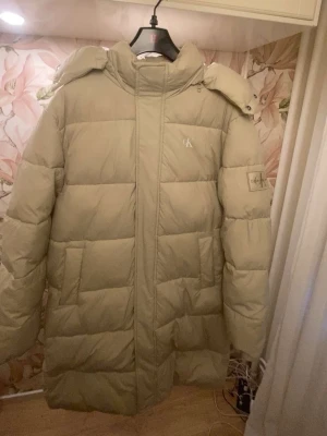 Beige lång pufferjacka från Calvin Klein - Säljer en beige lång parkajacka från Calvin Klein med luva som man kan ta av med hjälp av dragkedja. Jackan har diskret CK-logga på bröstet och ärmarna, två djupa sidofickor och dubbelsidig dragkedja. Väldigt varm och gosig och extremt bra på att hålla dig varm under viktern