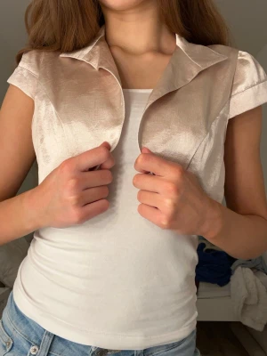 Champagnefärgad bolero i satin - Säljer en champagnefärgad, glansig bolero i satin-look. Boleron har en snygg krage och är ärmlös med en croppad passform. Perfekt att styla över ett linne eller klänning för en trendig touch. Perfekt för nyår och fester samt en extra touch till en tråkig outfit.