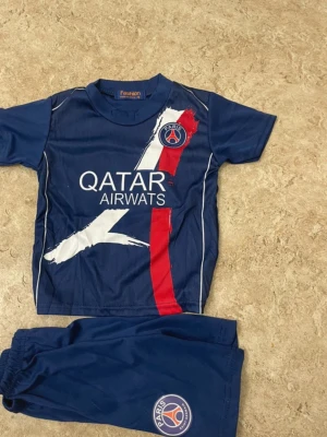 PSG fotbollströja O. Dembélé #10 - Säljer en blå PSG fotbollströja med O. Dembélé och nummer 10 på ryggen. Tröjan har tryck med Qatar Airways och klubbens emblem på bröstet, samt matchande blå shorts med PSG-logga. Materialet är lätt polyester, perfekt för fotbollsträning.