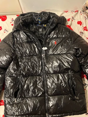 Svart pufferjacka från Polo Ralph Lauren - Säljer en svart, glansig pufferjacka från Polo Ralph Lauren med röd logga på bröstet. Jackan har huva, flera dragkedjeförsedda fickor och quiltad design. Perfekt för kalla dagar och har en riktigt snygg streetstil.