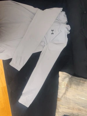 Ljusgrå vindjacka från Under Armour - Snygg ljusgrå vindjacka från Under Armour med svart logga på bröstet och tryck på ryggen. Jackan har huva med snörning, hel dragkedja och långa ärmar med resår vid muddar. Perfekt för träning och aktiva dagar utomhus.