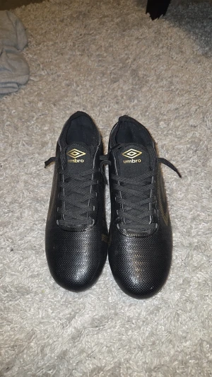 Svarta Umbro fotbollsskor med guld - Säljer ett par svarta fotbollsskor från Umbro med snygga gulddetaljer och logga på sidan och tungan. Skorna har snörning och gult mönster på dobbarna under sulan. Ovandelen är i syntetmaterial med diskret prickigt mönster för extra stil.