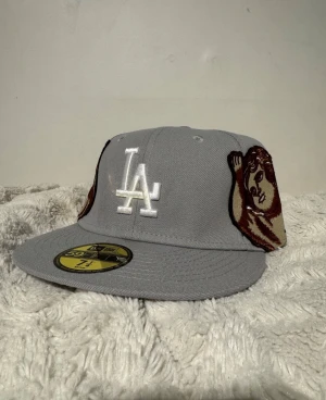Grå LA 59FIFTY keps med broderad ängel - Säljer en grå New Era 59FIFTY keps med LA-logga framtill och broderade änglar på sidorna. Baksidan har texten 'Ion Stan' i vit brodyr. Kepsen har platt skärm och klassisk passform. Perfekt för dig som vill sticka ut med coola detaljer och unik design.