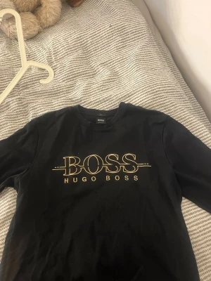 Svart långärmad t-shirt från Hugo Boss - Snygg svart långärmad t-shirt från Hugo Boss med stort guldigt BOSS-tryck på bröstet. Rund hals och klassisk passform. Perfekt för dig som gillar stilrena och exklusiva märken.