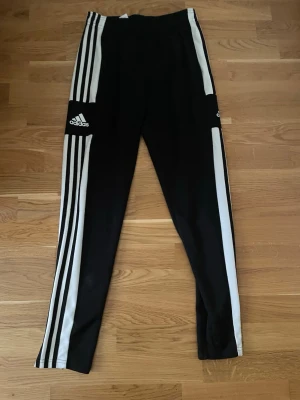 Svarta Adidas träningsbyxor med dragkedja - Svarta träningsbyxor från Adidas med vita ränder längs sidorna och logga på benet. Byxorna har dragkedja vid benslut och är gjorda i ett syntetmaterial som är perfekt för sport och träning. Snygg och sportig look med slim passform.