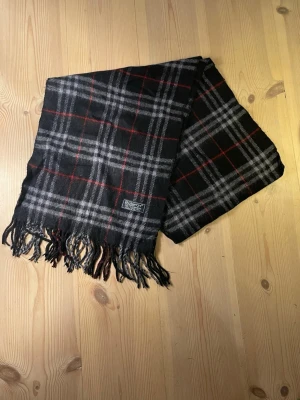 Rutig svart halsduk från Burberry - Snygg svart halsduk från Burberry med klassiskt rutigt mönster i grått och rött. Halsduken är gjord i mjuk ull och har fransar längs nederkanten. Perfekt för dig som gillar tidlös design och vill ha något som sticker ut lite extra.