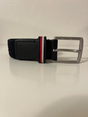 Svart flätad bälte från Tommy Hilfiger - Riktigt snyggt flätat bälte från Tommy Hilfiger i svart men är väldigt mörk blå igenterligen. Metall spänne och en detalj i rött, vitt och blått.