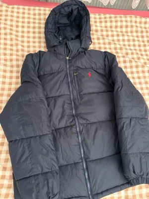 Mörkblå pufferjacka från Polo Ralph Lauren - Snygg mörkblå pufferjacka från Polo Ralph Lauren med huva och dragkedja framtill. Jackan har ett rött broderat Polo-logo på bröstet, praktiska fickor och justerbara muddar. Perfekt för kalla dagar och har en schysst oversized vibe.