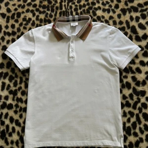 Burberry Piké - Snygg vit pikétröja från Burberry med klassisk krage i brunt, beige och svart rutigt mönster. Tröjan har korta ärmar och knäppning framtill. Perfekt för en clean och stilren look. Tröjan är i nyskick!  Priset kan diskuteras😇
