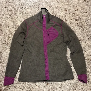 Grå tunn fleecejacka med lila detaljer från Craft - Sportig grå tunn fleecejacka från Craft i storlek L, med lila detaljer på dragkedja, armbågar, bröstficka och muddar. Jackan har hög krage, lång ärm och en bröstficka med dragkedja. Perfekt för lager-på-lager och utomhusaktiviteter. Koftan är lite smutsig på höger armbåge, men det går säkert bort i tvätten.