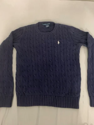 Mörkblå stickad tröja från Ralph Lauren - Säljer en mörkblå stickad tröja från Ralph Lauren i 100% bomull. Tröjan har kabelstickat mönster, rund hals och långärmade ribbade muddar. Klassisk vit Polo-logga broderad på bröstet. Perfekt för dig som gillar stilrena och tidlösa plagg.