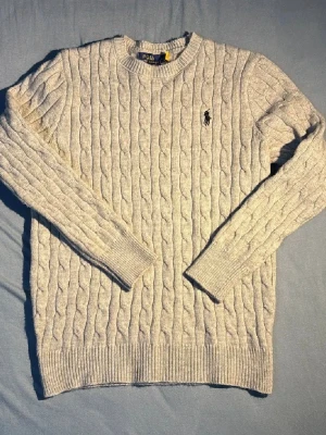 Grå kabelstickad tröja Polo Ralph Lauren - Säljer en grå kabelstickad tröja från Polo Ralph Lauren i storlek S. Tröjan har rund hals, ribbade muddar och det klassiska broderade logot på bröstet. Perfekt för dig som gillar stilrena och tidlösa plagg med snygg struktur.