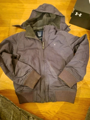 Grå vindjacka med huva från GANT - Grå vindjacka från GANT med huva och mjukt fleecefoder. Jackan har ribbade muddar vid ärmslut och nederkant, dragkedja framtill och två sidofickor. Perfekt för kyliga dagar och har en clean, sportig look.