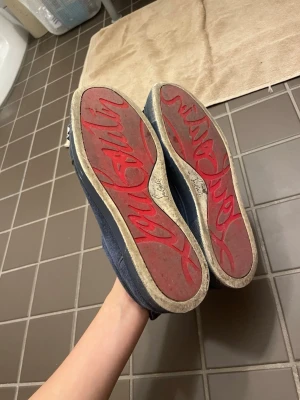 Köp inte jag byter - Säljer ett par blå sneakers från Christian Louboutin med ikoniska röda sulor och logotypen ingraverad undertill. Skorna har en klassisk rund tå och platt sula, perfekt för dig som vill sticka ut med exklusiv design och snygga detaljer.