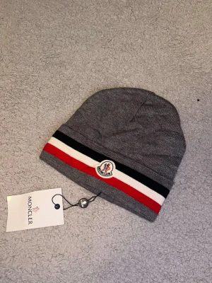 Grå Moncler mössa med ränder - Snygg grå Moncler mössa med klassiska svarta, vita och röda ränder nertill. Mössan har Moncler-logga framtill och är tillverkad i mjukt stickat material. Perfekt accessoar för att hålla stilen under kyliga dagar.Aktiv 24/7✅ Skickas inom 24h📦🚚 Kan gå ner i hugget 😃✅ Hör av er via frågor