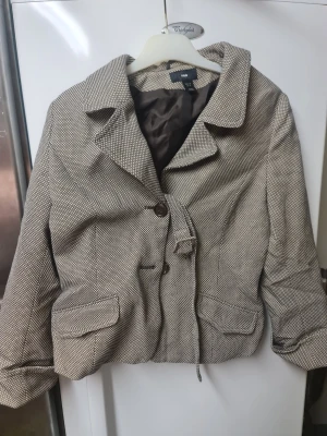 Rutig kavajjacka från H&M, stl 38 - Snygg kavajjacka från H&M i beige och brunt rutigt mönster. Klassisk krage, två knappar framtill och fickor med lock. Jackan är fodrad och har en croppad passform som ger en trendig look. Perfekt att slänga över en hoodie eller t-shirt.