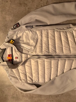 Ljusgrå dunväst från Parajumpers XL - Säljer en ljusgrå dunväst från Parajumpers i storlek XL. Västen har quiltad design, dragkedja framtill och en snygg patch på axeln. Tillverkad i syntetmaterial och dun, perfekt för lager-på-lager och streetwear. Flera praktiska fickor och coola detaljer. Passar de som bör L jätte bra 
