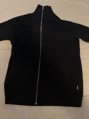 Svart stickad kofta med dragkedja - Säljer en svart stickad kofta med hel dragkedja framtill och hög krage. Koftan har två fickor på sidorna och en stilren, ribbad struktur. Perfekt för lager-på-lager och chill dagar. Passar dig som gillar enkel och clean stil.