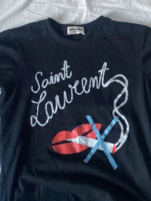 Svart Saint Laurent t-shirt - Svart t-shirt från Saint Laurent med tryck i vitt, rött och blått framtill. Motiv med texten 'Saint Laurent', röda läppar och grafiska detaljer. Klassisk rund hals och korta ärmar. Perfekt för en cool och avslappnad stil.