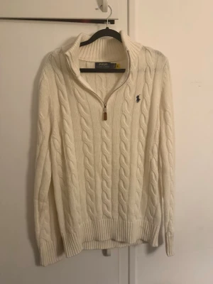 Vit stickad tröja från Polo Ralph Lauren - Säljer en vit kabelstickad tröja från Polo Ralph Lauren med half zip och ribbad krage. Tröjan har långärmad design, klassisk logga broderad på bröstet och grova muddar vid ärmslut och nederkant. Perfekt för kyliga dagar och snygg till jeans.
