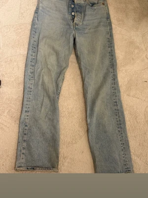 Levi's 501 ljusblå jeans straight fit - Ljusblå Levi's 501 jeans med klassisk straight fit och knappgylf. Snygga bakfickor med den ikoniska röda Levi's-taggen och läderpatch i midjan. Jeansen är tillverkade i slitstarkt bomullsjeansmaterial och har kontrastsömmar. Liten defekt på sista bilden men sytt ihop så syns inte mycket