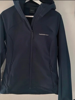 Mörkblå vindjacka Peak Performance - Snygg mörkblå vindjacka från Peak Performance med huva och dragkedja framtill. Jackan har två praktiska fickor med dragkedja och en stilren design. I storlek S. 
