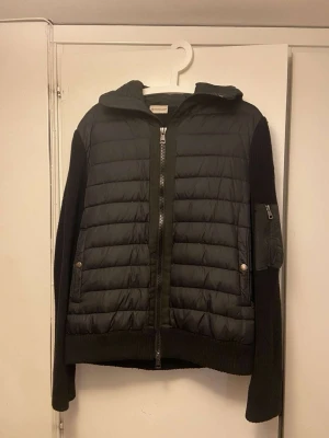 Svart pufferjacka från Moncler - Svart pufferjacka från Moncler med stickade ärmar och huva. Jackan har quiltad framsida, dragkedja och fickor med tryckknappar. Ärmarna har en snygg fickdetalj med dragkedja. Perfekt för dig som gillar street och vill ha något som sticker ut.
