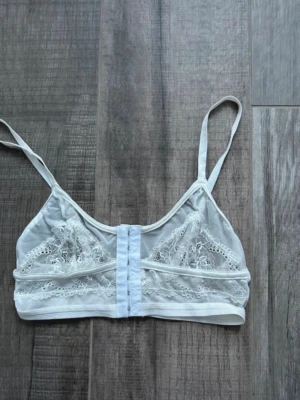Vit spetsdetaljerad bralette - Säljer en vit bralette med tunna axelband och vackra spetsdetaljer framtill. Modellen har hakknäppning fram och är gjord i ett mjukt, transparent material med broderade blommor i spetsen. Perfekt för dig som gillar en feminin och stilren look.
