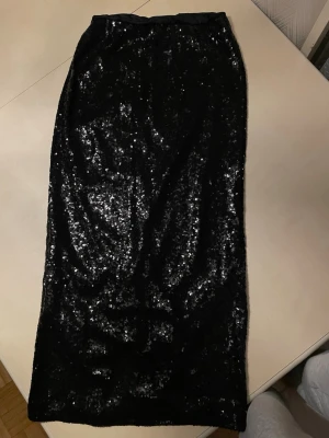 Svart paljettkjol från Gina Tricot - Säljer en svart långkjol från Gina Tricot, täckt med glittriga paljetter som ger en riktigt snygg och festlig look. Kjolen har en slits bak för extra rörelsefrihet och är fodrad med mjukt tyg på insidan. Perfekt för dig som vill sticka ut.